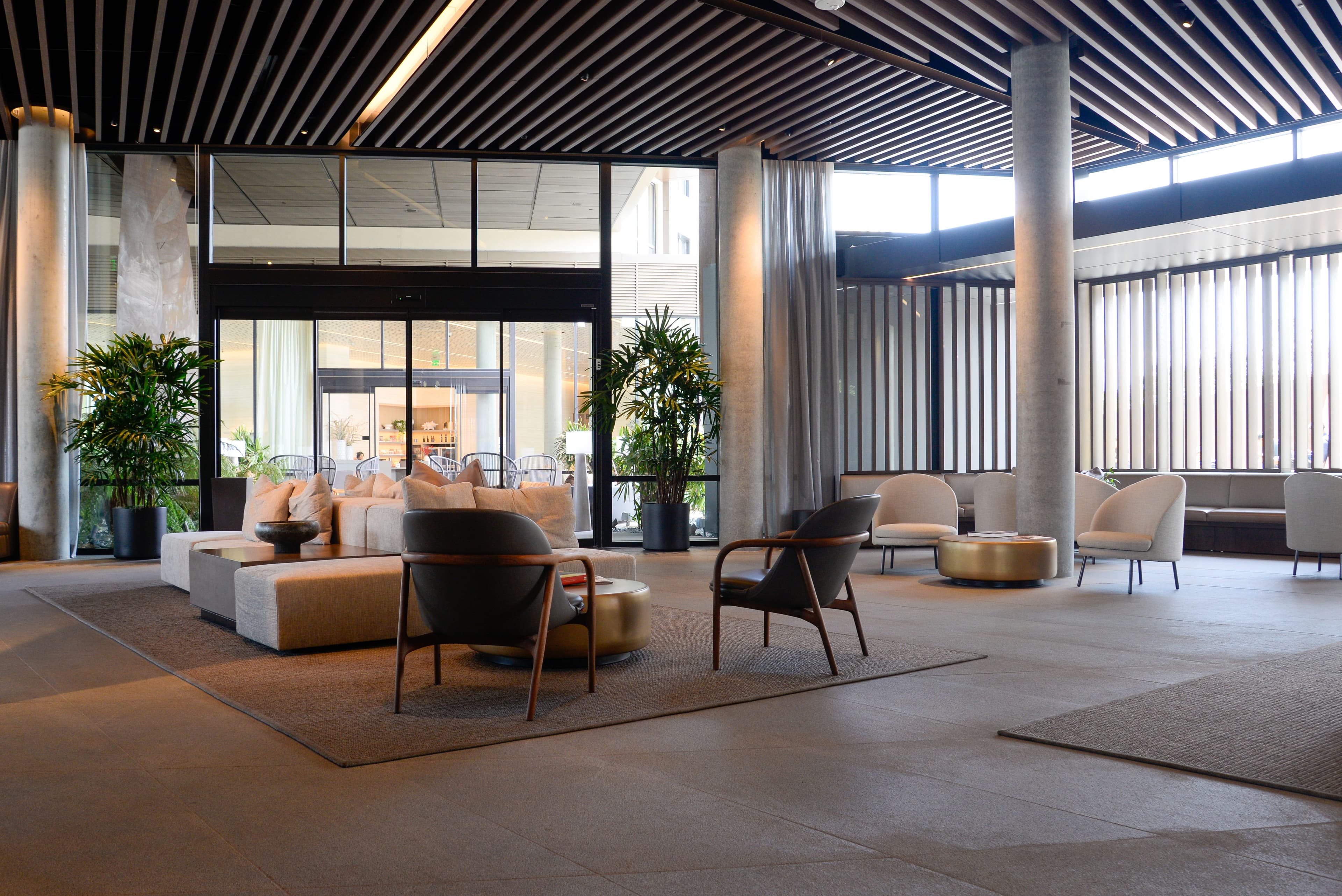 Modern lobby with neutral furniture, gold coffee tables, potted plants, and a slatted wood ceiling.