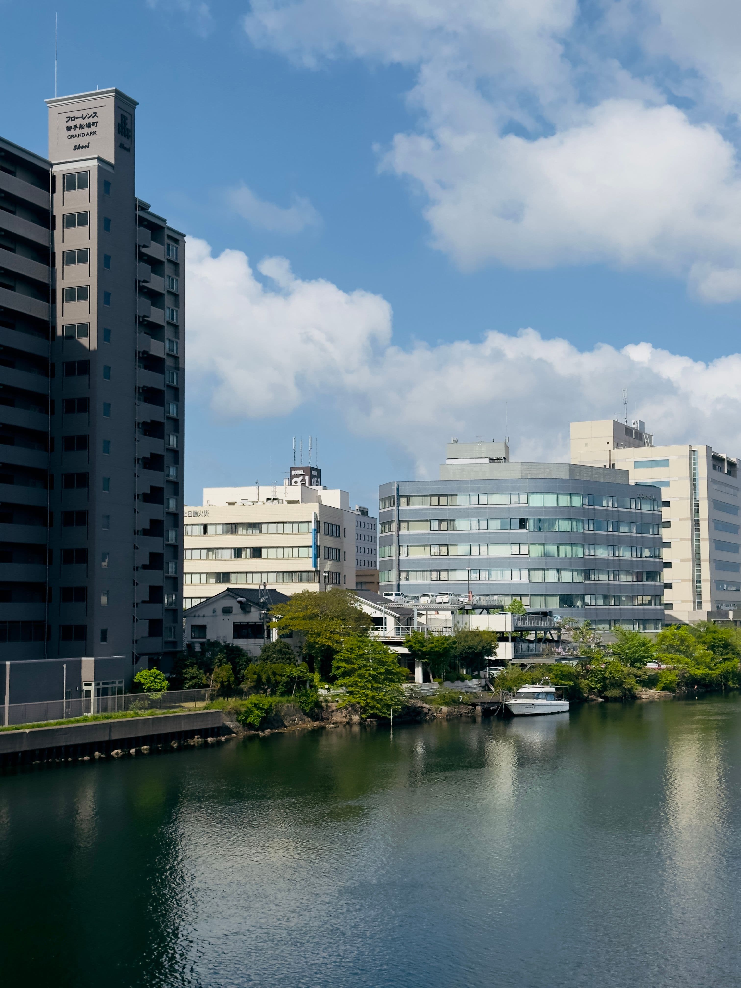 Modern buildings and trees line a calm river reflecting a bright, cloudy blue sky.
