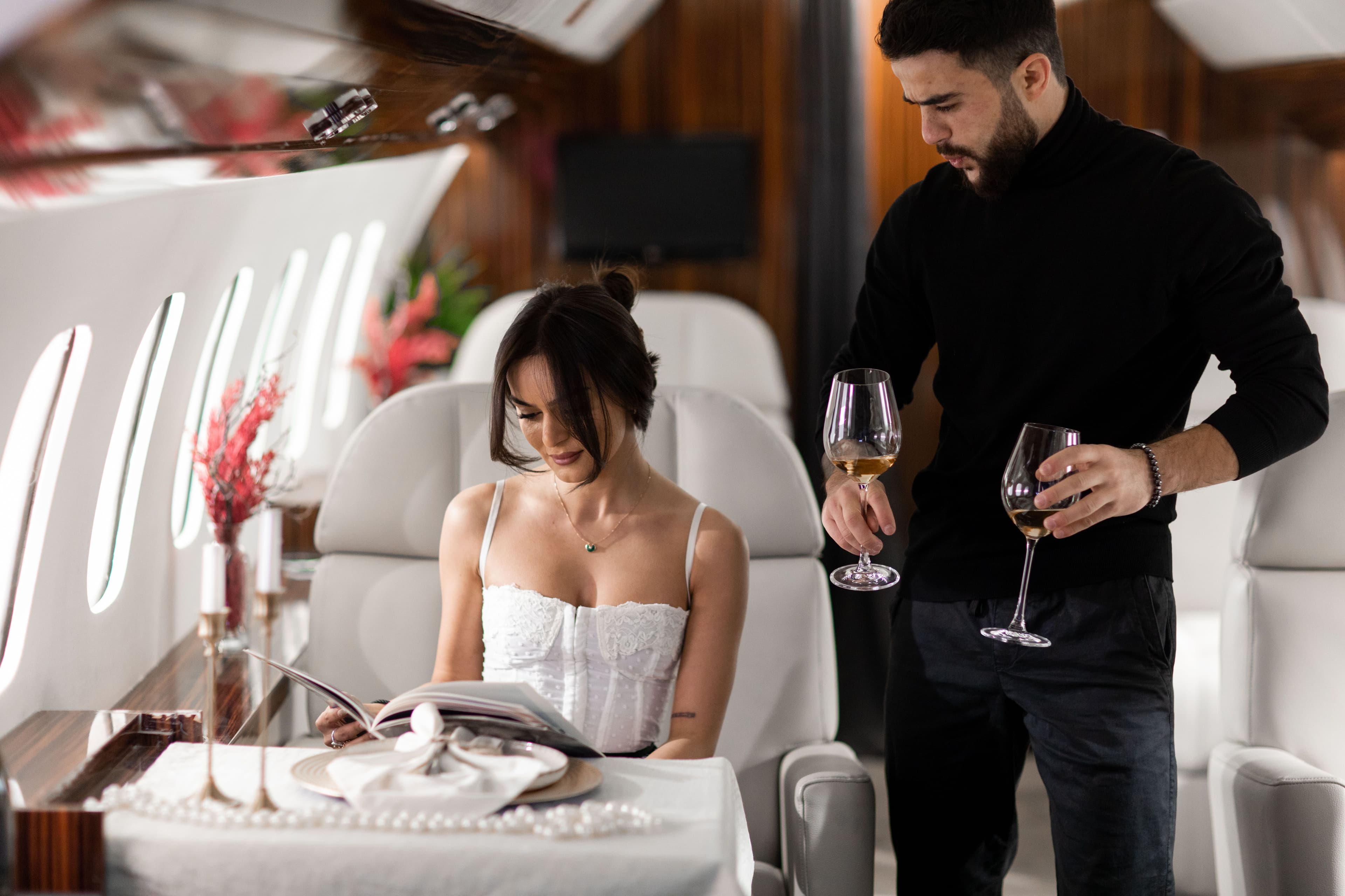 A man serves wine to a woman reading a magazine inside a luxury private jet.